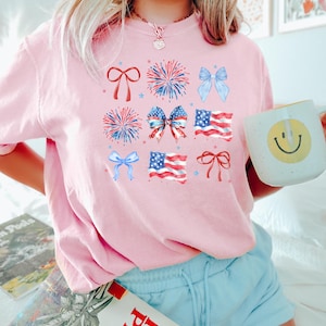 May include: A pink t-shirt with a patriotic design featuring red, white, and blue stars, fireworks, bows, and American flags.