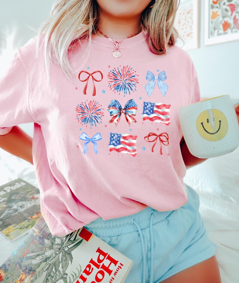 May include: A pink t-shirt with a patriotic design featuring red, white, and blue stars, fireworks, bows, and American flags.