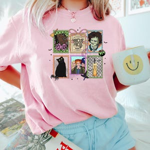Puede incluir: Camiseta rosa claro de cuello redondo con un gr&aacute;fico colorido de dise&ntilde;os de sellos postales con temas de Halloween. Los sellos incluyen un gato negro, un zombi y el texto "Hocus Pocus". La camiseta est&aacute; hecha de un material suave.
