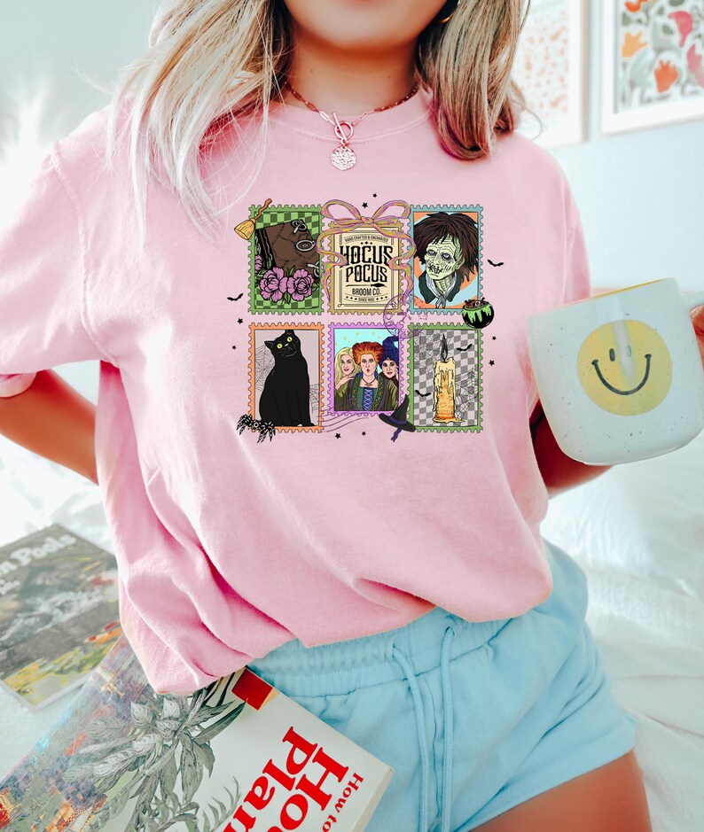 Puede incluir: Camiseta rosa claro de cuello redondo con un gr&aacute;fico colorido de dise&ntilde;os de sellos postales con temas de Halloween. Los sellos incluyen un gato negro, un zombi y el texto "Hocus Pocus". La camiseta est&aacute; hecha de un material suave.