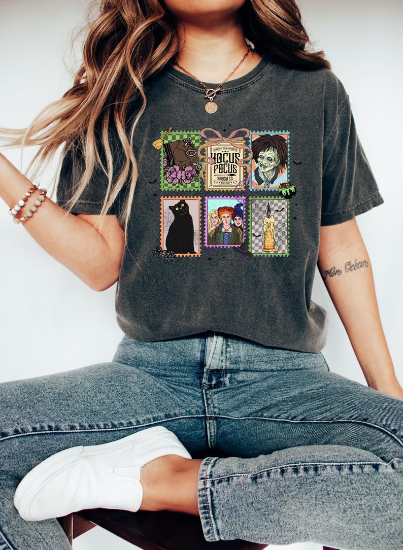 Puede incluir: Camiseta gris oscuro con un gr&aacute;fico colorido de sellos postales con tem&aacute;tica de Hocus Pocus. Los sellos representan un gato negro, un caballo, un zombi, las hermanas Sanderson, una vela y las palabras "Hocus Pocus Broom Co."