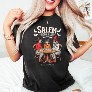 Puede incluir: Camiseta negra con un gr&aacute;fico de tres figuras sentadas alrededor de una mesa, con el texto "Salem Book Club - 1692" y "Massachusetts EST. 1692". La ilustraci&oacute;n presenta un diablo, una ni&ntilde;a y un esqueleto con un sombrero de bruja.