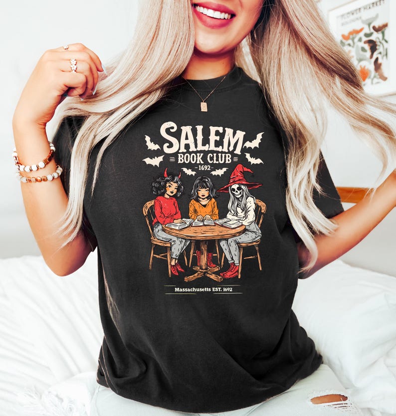 Puede incluir: Camiseta negra con un gr&aacute;fico de tres figuras sentadas alrededor de una mesa, con el texto "Salem Book Club - 1692" y "Massachusetts EST. 1692". La ilustraci&oacute;n presenta un diablo, una ni&ntilde;a y un esqueleto con un sombrero de bruja.