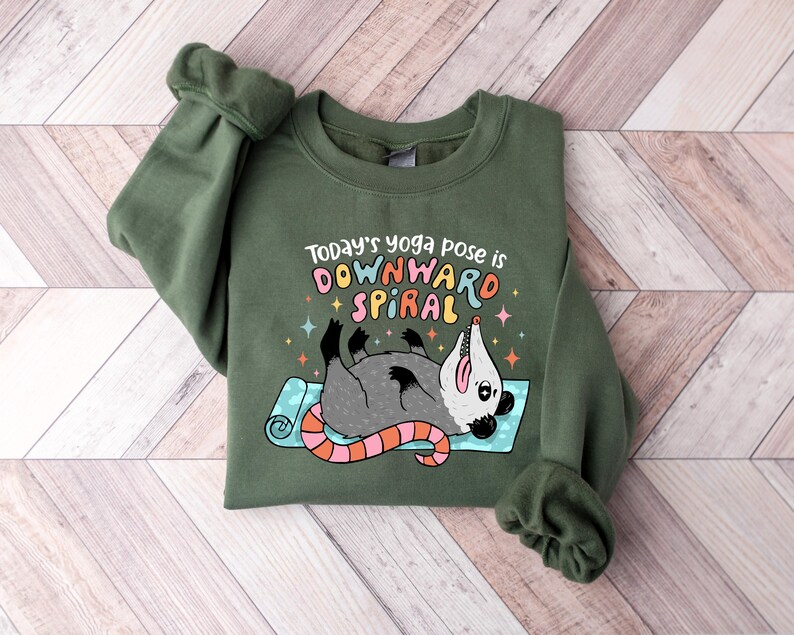 May include: Green sweatshirt with the text "Today's yoga pose is DOWNWARD SPIRAL" and a cartoon opossum lying on a yoga mat. The opossum is gray with a pink nose and tail. The text is in multiple colors.