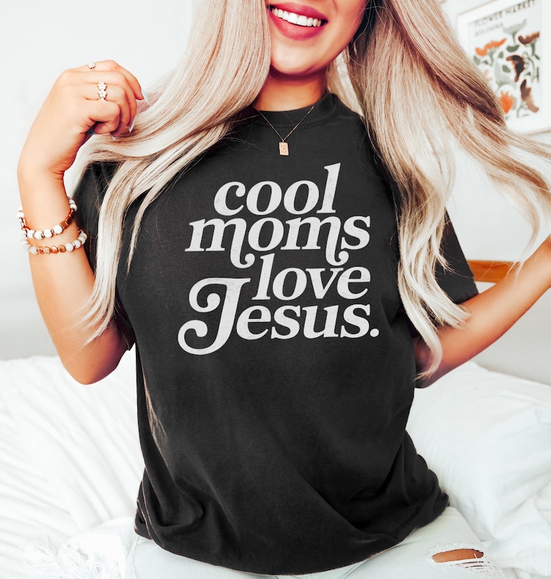 May include: Black t-shirt with the white text "cool moms love Jesus." The shirt is a crew neck style and appears to be made of a soft material. The text is in a bold, stylized font.