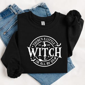 Op de afbeelding: Zwart sweatshirt met ronde hals en een witte cirkelvormige afbeelding met de tekst "There's a Little Witch in All of Us". Het ontwerp bevat een heks op een bezem, vleermuizen en het jaar 1692. Het sweatshirt ligt op een spijkerbroek.