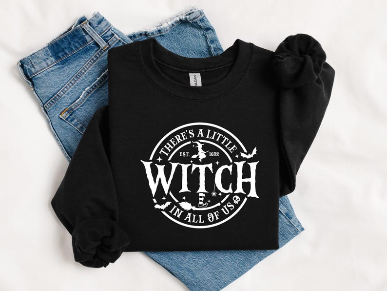 Op de afbeelding: Zwart sweatshirt met ronde hals en een witte cirkelvormige afbeelding met de tekst "There's a Little Witch in All of Us". Het ontwerp bevat een heks op een bezem, vleermuizen en het jaar 1692. Het sweatshirt ligt op een spijkerbroek.