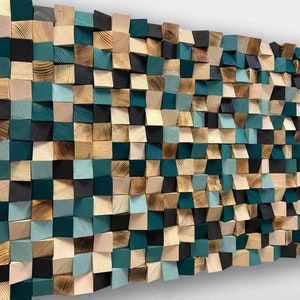 Op de afbeelding: Een wandkunstwerk bestaande uit houten blokken in verschillende tinten teal, zwart en natuurlijke houttinten. De blokken zijn gerangschikt in een mozaïekpatroon, waardoor een getextureerd, driedimensionaal effect ontstaat. Het kunstwerk is op een witte muur gemonteerd.