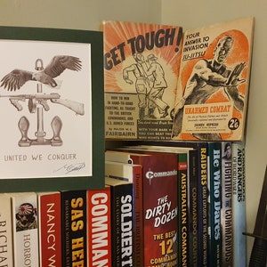 May include: A framed print featuring an eagle and a machine gun, with the words "UNITED WE CONQUER". A collection of vintage books and a combat knife are also present. The books include titles such as "Soldier" and "Commando".