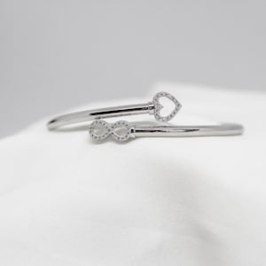 Puede incluir: Un brazalete plateado con diseño abierto. Un extremo presenta un diseño en forma de corazón, y el otro un símbolo de infinito, ambos incrustados con pequeñas piedras transparentes. El brazalete se muestra sobre una superficie blanca.