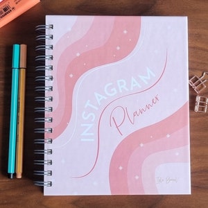 Op de afbeelding: Een roze en wit spiraalnotitieblok met de tekst "INSTAGRAM PLANNER" in witte letters. De omslag heeft een roze en wit golvend ontwerp met witte sterren.