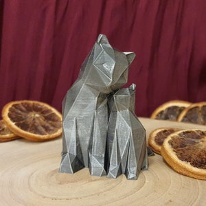 Könnte beinhalten: Zwei graue, geometrisch geformte Katzenfiguren, eine größer als die andere, stehen nebeneinander auf einer Holzoberfläche. Die größere Katze schaut die kleinere an, die nach vorne blickt. Die Figuren sind aus einem glatten, metallischen Material gefertigt.