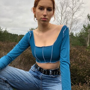 May include: A blue long-sleeved crop top with a square neckline and seams along the front. The top is worn with a black belt and blue jeans.