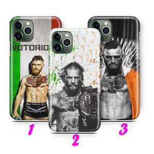 Puede incluir: Tres fundas de teléfono diferentes con una imagen en blanco y negro de un hombre con tatuajes y el texto "McGregor" y "UFC". La primera funda tiene un fondo verde, blanco y naranja con el texto "Notorio". La segunda funda tiene un fondo blanco con un patrón grunge en blanco y negro. La tercera funda tiene un fondo negro con un patrón gris y blanco.