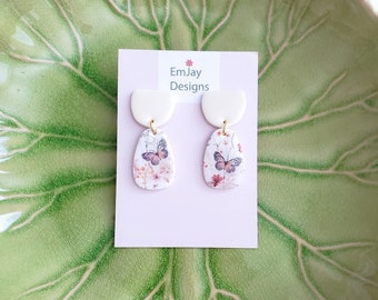 May include: A pair of white ceramic earrings with a floral and butterfly design. The earrings are on a pink card with the text "EmJay Designs".