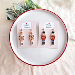 Könnte beinhalten: Zwei Paar Ohrhänger mit einem Nussknackersoldaten-Design. Ein Paar ist rosa und das andere rot, schwarz und blau. Jeder Ohrring hat eine goldfarbene Oberseite und wird auf einer rosa Karte mit dem Text "EmJay Designs" präsentiert.
