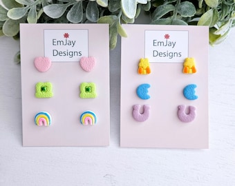 May include: Two sets of colorful polymer clay earrings on white cards. The first set features a pink heart, green four-leaf clover, and a rainbow. The second set features a yellow bear, a blue crescent moon, and a purple horseshoe.