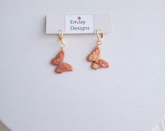 May include: A pair of gold hoop earrings with a pink butterfly charm. The butterfly charm has a gold leaf design. The earrings are on a white background with a card that says "EmJay Designs".