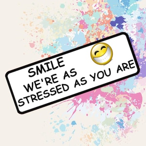 May include: A rectangular sign with a black border and the words "SMILE WE'RE AS STRESSED AS YOU ARE" in bold black font. A yellow smiley face with a wide grin is in the upper right corner. The background features colorful paint splatters.