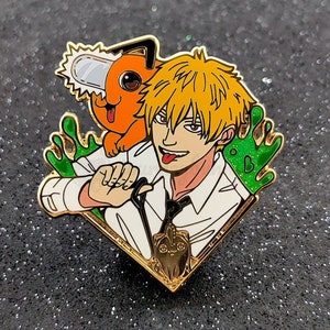 May include: Gold-coloured enamel pin featuring a cartoon character with orange fur and a chainsaw, a man with blond hair and a white shirt, and green slime. The man is holding a black and white cat.