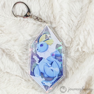 May include: A blue and yellow Pokemon character, Vaporeon, is encased in a clear iridescent prism-shaped keychain. The keychain is made of acrylic and has a silver metal ring.