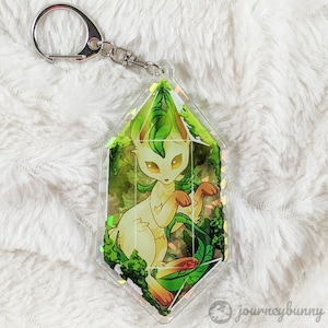 May include: A white and green crystal-shaped keychain with a cartoon fox inside. The fox is surrounded by green leaves and vines. The keychain is made of clear acrylic and has a silver key ring.
