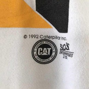 Może przedstawiać: Zbliżenie białej tkaniny z czarno-ż&oacute;łtym wzorem. Obraz zawiera tekst "&copy; 1992 Caterpillar Inc." oraz okrągłe logo ze słowem "CAT" w środku. Słowa "LICENSED PRODUCTS OF CATERPILLAR INC." otaczają logo.