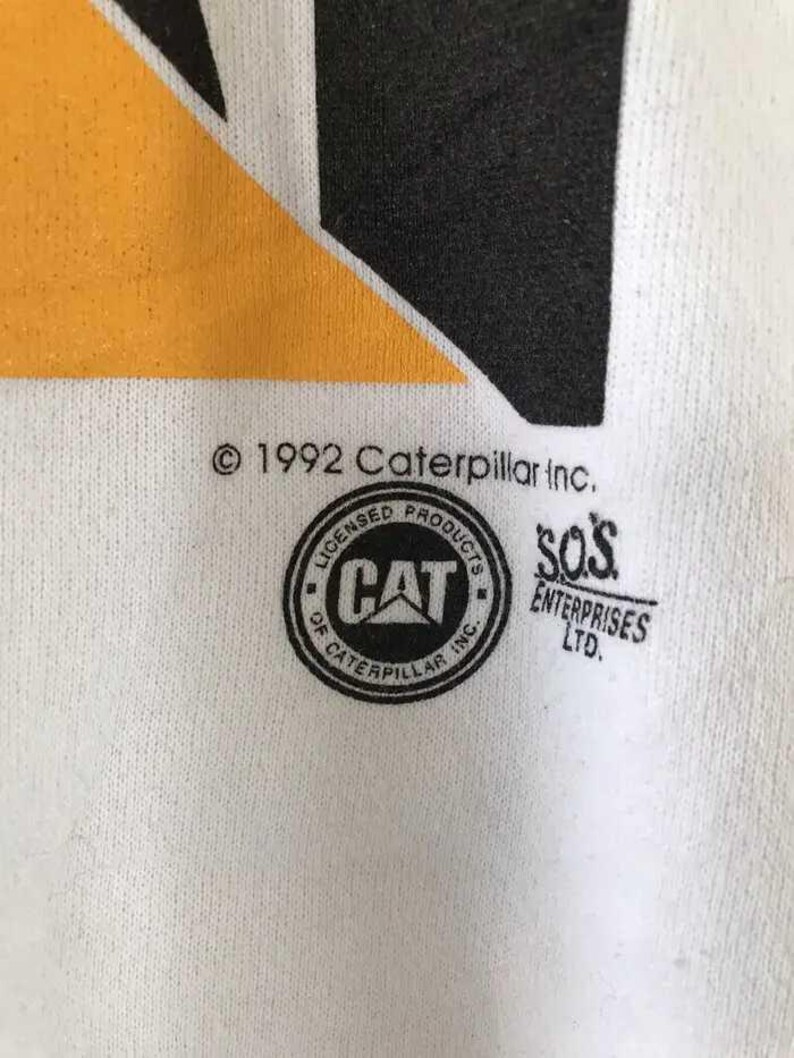 Może przedstawiać: Zbliżenie białej tkaniny z czarno-ż&oacute;łtym wzorem. Obraz zawiera tekst "&copy; 1992 Caterpillar Inc." oraz okrągłe logo ze słowem "CAT" w środku. Słowa "LICENSED PRODUCTS OF CATERPILLAR INC." otaczają logo.