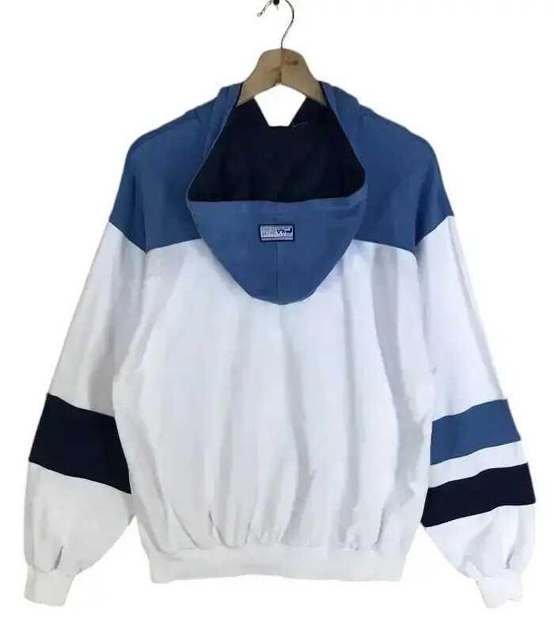Puede incluir: Chaqueta con capucha blanca, azul y azul marino colgada de una percha de madera. La chaqueta tiene un dise&ntilde;o de bloques de color con capucha y mangas azules, cuerpo blanco y rayas azul marino y azules en las mangas.