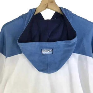 Puede incluir: Una sudadera con capucha de bloques de color azul, blanco y azul marino. La capucha es azul con el interior azul marino. El cuerpo de la sudadera con capucha es blanco. El logotipo de Fila est&aacute; en la capucha. La sudadera con capucha est&aacute; colgada en una percha de madera.