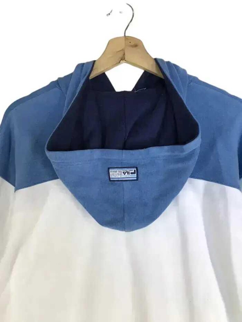 Puede incluir: Una sudadera con capucha de bloques de color azul, blanco y azul marino. La capucha es azul con el interior azul marino. El cuerpo de la sudadera con capucha es blanco. El logotipo de Fila est&aacute; en la capucha. La sudadera con capucha est&aacute; colgada en una percha de madera.