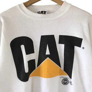 Może przedstawiać: Biała bluza z czarnym logo "CAT" i ż&oacute;łtym tr&oacute;jkątem. Bluza ma okrągły dekolt i długie rękawy. Logo zawiera nazwę marki Caterpillar i informację o prawach autorskich.