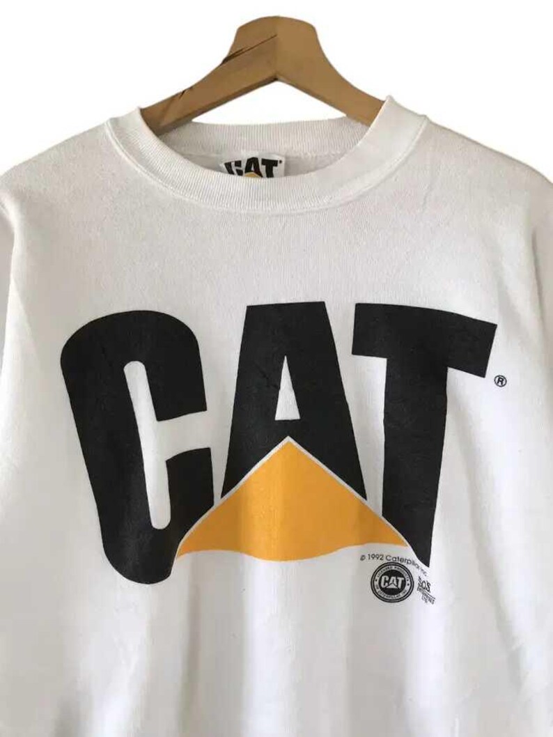 Może przedstawiać: Biała bluza z czarnym logo "CAT" i ż&oacute;łtym tr&oacute;jkątem. Bluza ma okrągły dekolt i długie rękawy. Logo zawiera nazwę marki Caterpillar i informację o prawach autorskich.