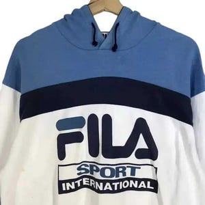 Puede incluir: Una sudadera con capucha Fila azul, blanca y azul marino. La parte superior de la sudadera con capucha es azul, con una franja azul marino en el pecho. La parte inferior es blanca, con el logotipo de Fila y "SPORT INTERNATIONAL" impreso.