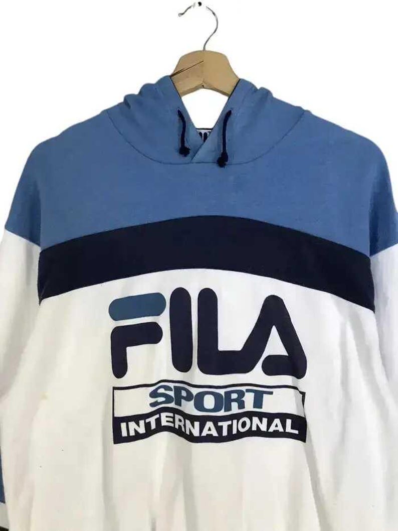 Puede incluir: Una sudadera con capucha Fila azul, blanca y azul marino. La parte superior de la sudadera con capucha es azul, con una franja azul marino en el pecho. La parte inferior es blanca, con el logotipo de Fila y "SPORT INTERNATIONAL" impreso.