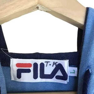 Puede incluir: Primer plano de la etiqueta de una camisa Fila azul y azul marino. La etiqueta es blanca con el logotipo de Fila en azul marino y rojo. La camisa est&aacute; colgada en una percha de madera.