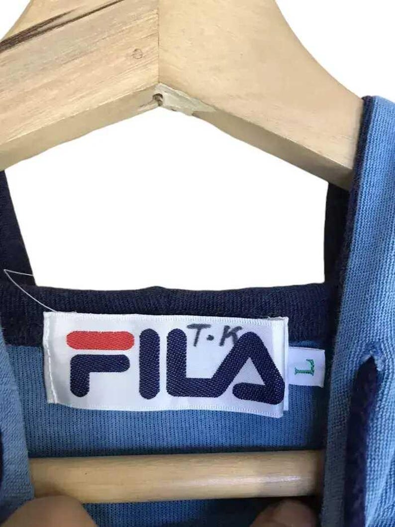 Puede incluir: Primer plano de la etiqueta de una camisa Fila azul y azul marino. La etiqueta es blanca con el logotipo de Fila en azul marino y rojo. La camisa est&aacute; colgada en una percha de madera.