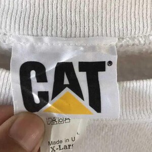 Może przedstawiać: Biała metka bluzy z czarnymi literami "CAT" nad ż&oacute;łtym tr&oacute;jkątem. Etykieta zawiera r&oacute;wnież instrukcje prania i tekst "Made in U. X-Large".