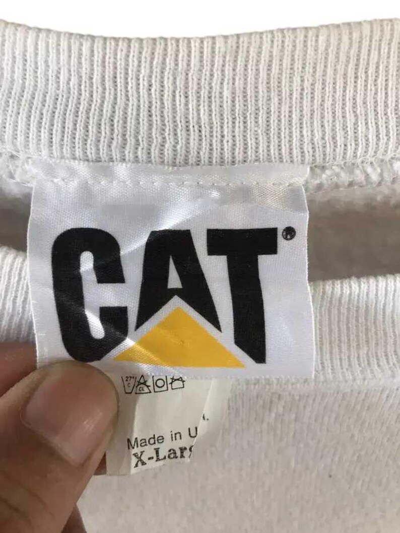 Może przedstawiać: Biała metka bluzy z czarnymi literami "CAT" nad ż&oacute;łtym tr&oacute;jkątem. Etykieta zawiera r&oacute;wnież instrukcje prania i tekst "Made in U. X-Large".