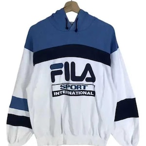 Puede incluir: Sudadera con capucha FILA Sport International blanca, azul y azul marino. La sudadera tiene una capucha y mangas azules, con un cuerpo blanco. El logotipo de FILA y el texto "SPORT INTERNATIONAL" est&aacute;n impresos en la parte delantera.