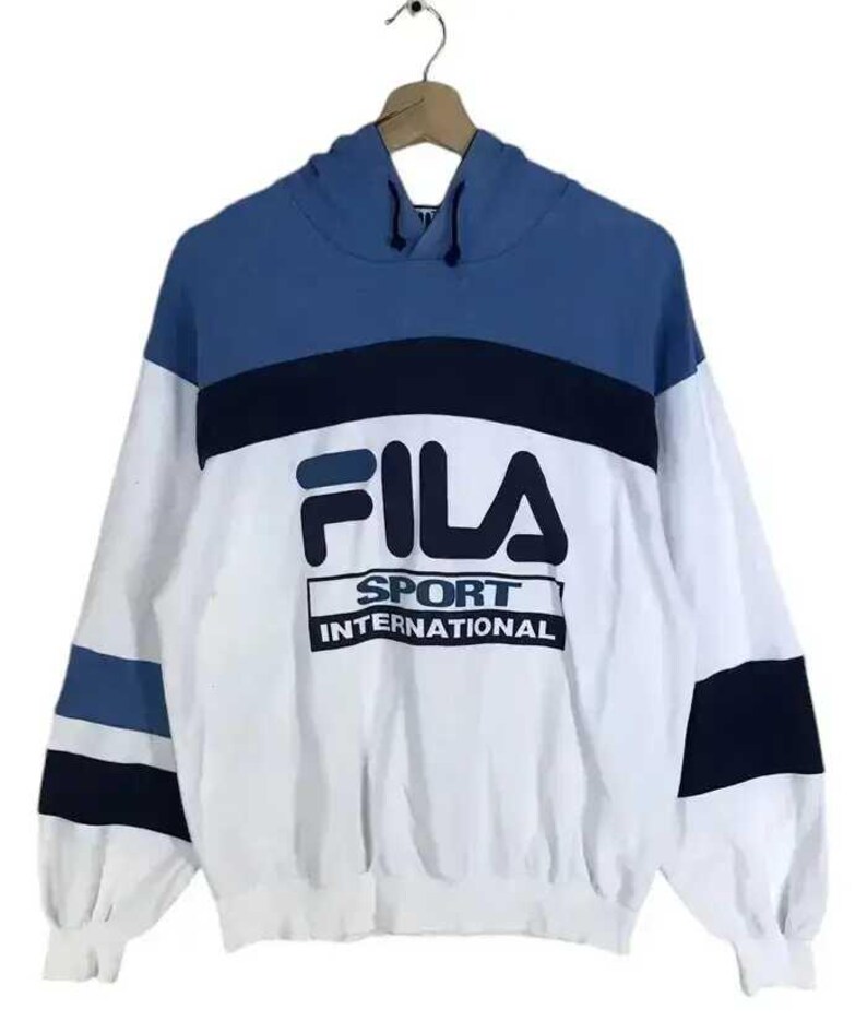 Puede incluir: Sudadera con capucha FILA Sport International blanca, azul y azul marino. La sudadera tiene una capucha y mangas azules, con un cuerpo blanco. El logotipo de FILA y el texto "SPORT INTERNATIONAL" est&aacute;n impresos en la parte delantera.