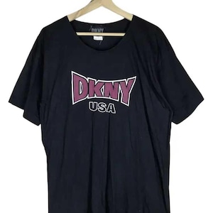 May include: Black DKNY USA t-shirt with a crew neck. The shirt features a maroon and white DKNY logo across the chest. The shirt is hanging on a wooden hanger.