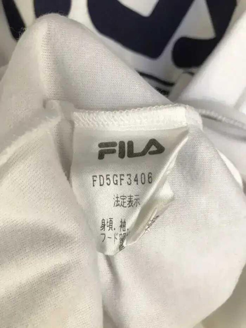 Puede incluir: Primer plano de una etiqueta de prenda blanca con el logotipo de FILA en gris. La etiqueta muestra el texto "FD5GF3406" y caracteres japoneses. La prenda parece estar hecha de una tela blanca suave.