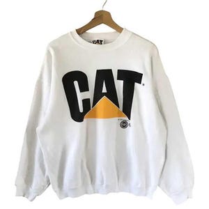 Może przedstawiać: Biała bluza z okrągłym dekoltem z napisem "CAT" dużymi czarnymi literami, ż&oacute;łtym tr&oacute;jkątem i małym logo. Bluza wisi na drewnianym wieszaku.