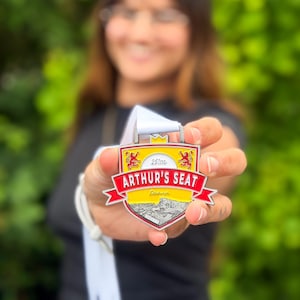 May include: A shield-shaped metal medal with a white ribbon. The design features a yellow background, red borders, and two red lions. It displays the text '251m', 'ARTHUR'S SEAT', and 'Edinburgh' above a black and white illustration of a hill.