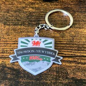 Może przedstawiać: Srebrny brelok do kluczy z zielono-czerwono-białym breloczkiem w kształcie tarczy. Brelok przedstawia walijskiego smoka, napis "Snowdon Yr Wyddfa" i liczbę "1085m".