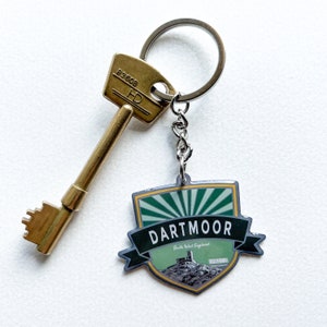 Op de afbeelding: Een messing sleutel en een sleutelhanger met een Dartmoor-ontwerp. De sleutelhanger heeft een schildvorm met een groen en wit ontwerp, het woord "DARTMOOR" in het wit en de tekst "South West England".