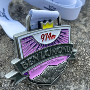 Puede incluir: Una medalla de plata con un diseño morado, rosa y blanco. Presenta el texto "Ben Lomond" y "974m" con una corona en la parte superior. La medalla también incluye el texto "Loch Lomond & The Trossachs".