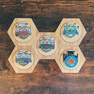 Puede incluir: Seis placas hexagonales de madera con medallones de metal que presentan diferentes diseños y textos. Las placas están dispuestas en forma de panal. Los medallones presentan textos como "Snowdonia 714m", "Yorkshire Three Peaks", "Muro de Adriano 135 km" y "Edale Skyline 32 km".
