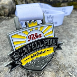 May include: A silver medal with a yellow and black design. The medal features a sun with rays, the text "978m Scafell Pike", and the words "Lake District" in a yellow banner.